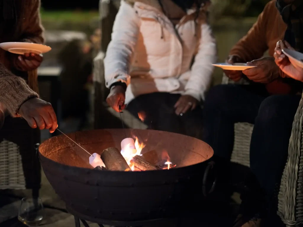Several people in coats roasting marshmallows over an outdoor fire pit.